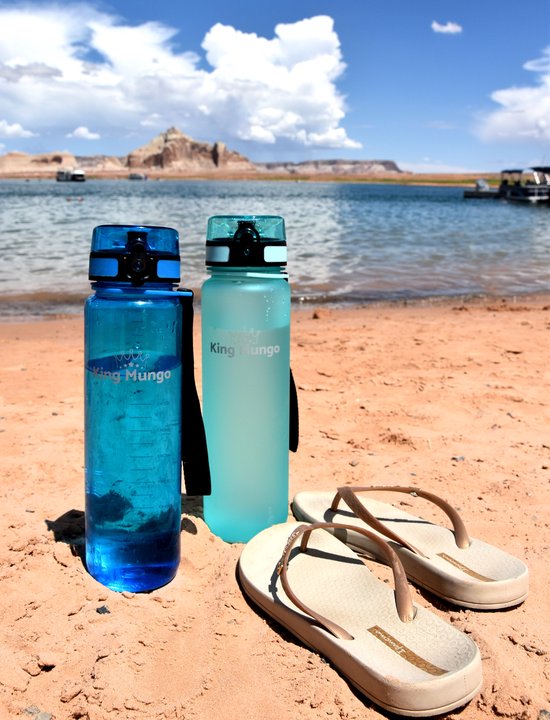 Kleine blauwe en lichtblauwe King Mungo drinkflessen op een strand met teenslippers.