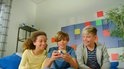Drie kinderen die op een bank zitten en lachen, terwijl de middelste jongen een gekleurde kubus vasthoudt.