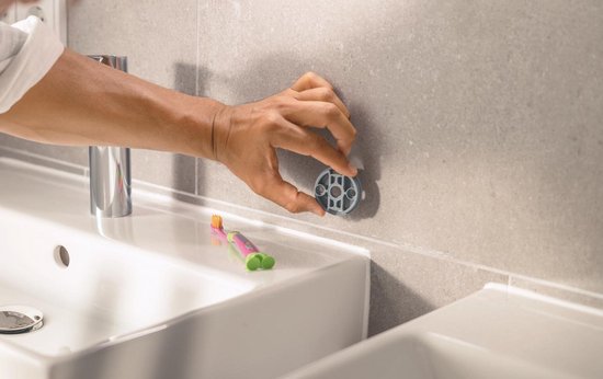 Une main fixe un support mural gris sur un mur carrelé gris, à côté d'un lavabo.