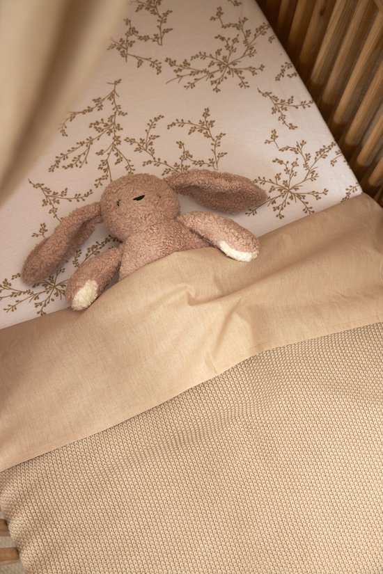 Een bruine konijnenknuffel ligt op een bedje in een wieg met een laken met bladerprint.