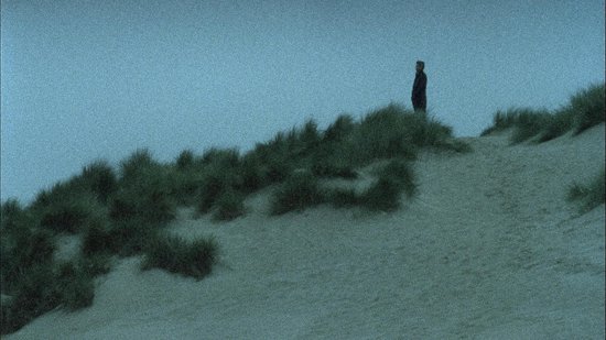Une personne se tient sur une dune herbeuse, sur un fond de ciel bleu-gris.