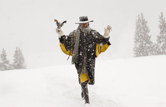 Man met zwarte hoed, sjaal en jas met gele accenten, houdt revolvers vast in de sneeuw.
