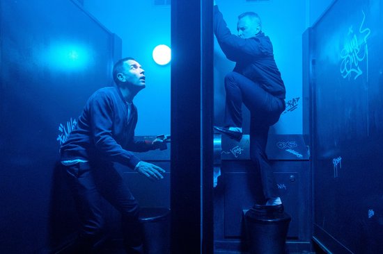 Twee mannen in een blauw verlichte badkamer met graffiti, één kijkt omhoog, de ander klimt.