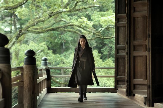 Shu Qi in een lang zwart gewaad en laarzen loopt op een houten pad onder een groene boomgaard.