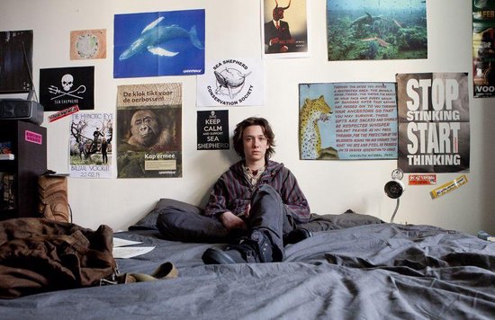 Jonge man zit op bed in kamer vol met posters, waaronder 'STOP STINKING START THINKING'.