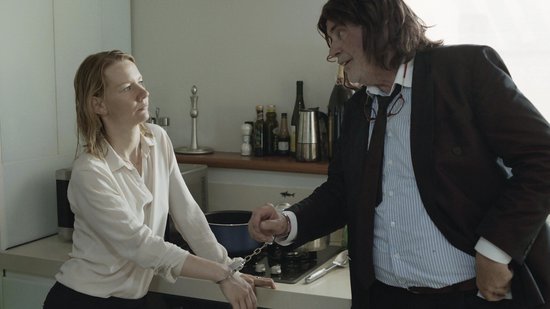 Een vrouw met een witte blouse staat met handboeien om haar polsen, tegenover een man in een pak met een rode stropdas.