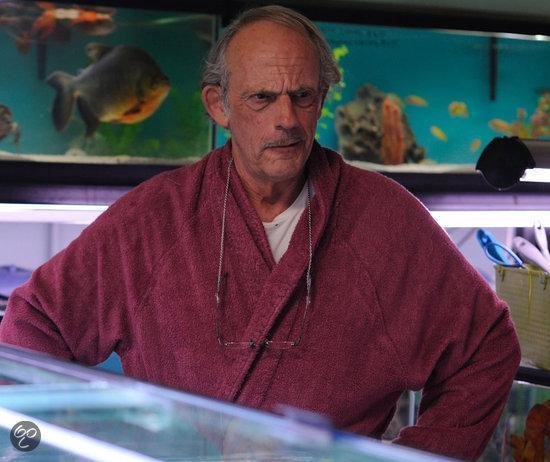 Christopher Lloyd met snor draagt een bordeauxrode badjas en een bril op een ketting.