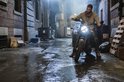 Een man in een beige trui en spijkerbroek zit op een motorfiets in een donker steegje met natte bestrating.