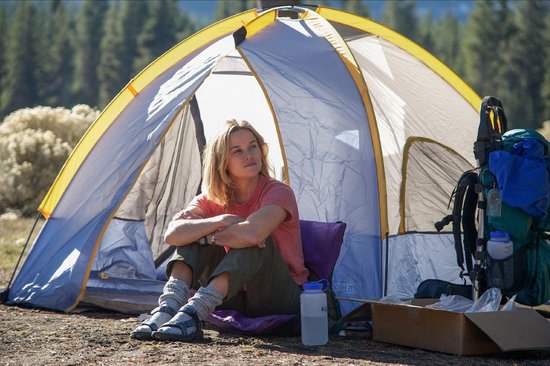 Reese Witherspoon assise dans une tente, vêtue d'un t-shirt orange et d'une écharpe à carreaux autour du cou. Un sac à dos est visible en arrière-plan.