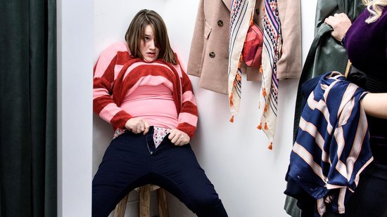 Vrouw in een pashokje met gestreepte trui en met moeite haar broek sluitend.
