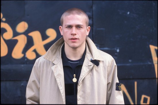 Charlie Hunnam met een geschoren hoofd, een beige trenchcoat en een zwarte jas, met een medaillon om zijn nek.