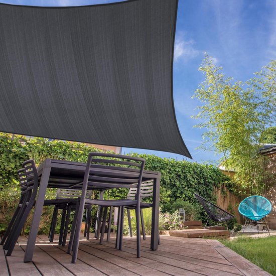Donkergrijs vierkant schaduwdoek hangt boven een tuintafel en stoelen op een houten terras, met begroeiing en een blauwe stoel op de achtergrond.