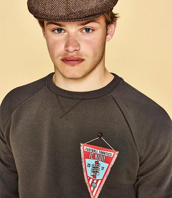 Jeune homme portant une casquette et un pull foncé avec un écusson rouge et bleu appliqué.