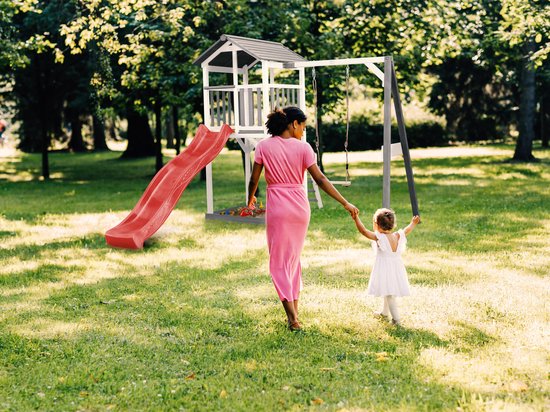 Vrouw in roze jurk en meisje in witte jurk lopen hand in hand in de tuin bij een speeltoestel.