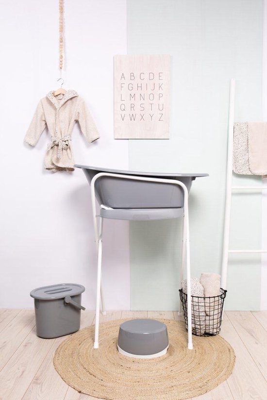 Beige badjas met stippen, grijs babybadje op een standaard, mand met handdoeken, emmer en een opstapje.