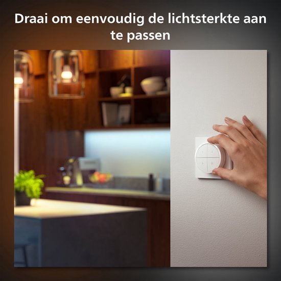 Een hand draait aan de witte Philips Hue Tap Dial Switch, die op een muur is bevestigd, om de lichtsterkte aan te passen. Op de achtergrond is een keuken te zien.