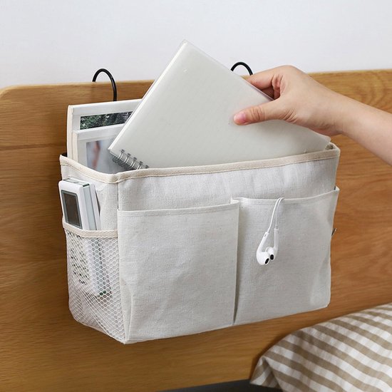 Witte stoffen bedorganizer met haken, hangend aan een houten bedrand. Een hand plaatst een boek in de organizer, met een koptelefoon aan de zijkant.