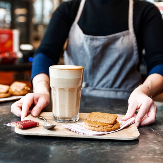 Barista met schort serveert glas met koffie en twee Biscoff koeken met crème ertussen.