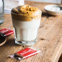 Glaasje met witte koffie met schuim en gemalen koekjes, met twee verpakte Lotus Biscoff koekjes.