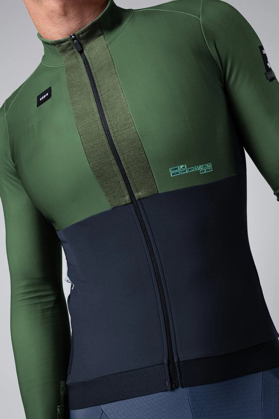 Model draagt een groen en donkerblauw Gobik fietsjack met lange mouwen, met rits en horizontale banden.