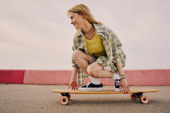 Een vrouw met een geel shirt, bruine short, witte sokken en sneakers, zittend op een longboard.