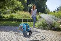 Vrouw sproeit planten met een slang vanuit een blauwe haspel op een betegeld terras.