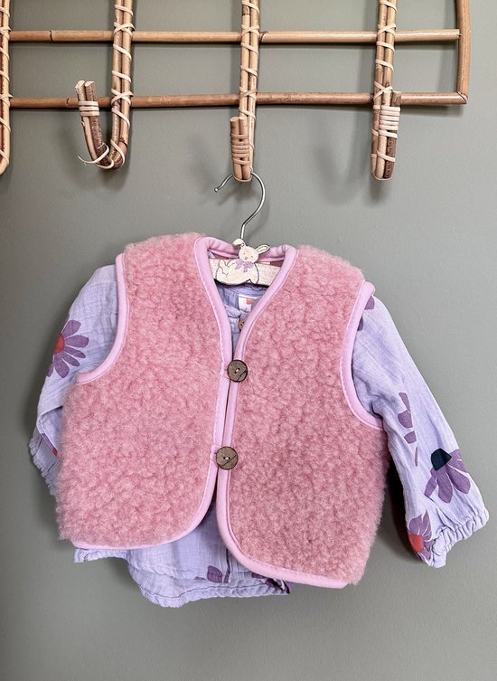 Roze wollen gilet met bruine knopen over een paars bloemenprint shirtje, hangend aan een kapstok.