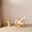 Schommelstoel met licht houten frame, crèmekleurige bouclé bekleding en een bijpassend kussen. Naast de stoel staat een gestreepte bijzettafel met vaas.