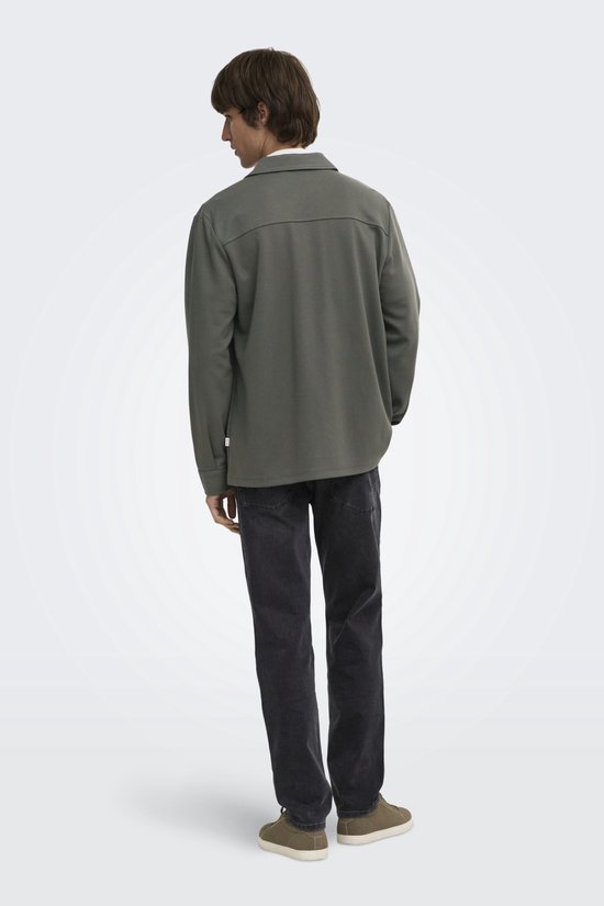 Man met de rug naar de camera, draagt olijfgroene utility blouse, grijze jeans en bruine sneakers.