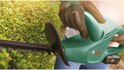 Een close-up van een Bosch heggenschaar die een groene heg snoeit, met pijlen die de snijrichting aangeven.