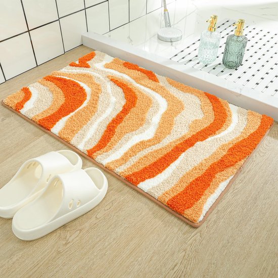 Badmat met golvend patroon in oranje, crème en wit, naast witte slippers.