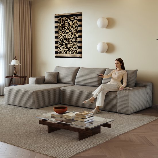 Grijze ribfluwelen hoekbank met chaise longue, bezet door een vrouw met een afstandsbediening.