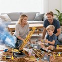 Gezin speelt op zand met een gele bouwkraan. De kraan hijst een grijze container. De ouders kijken lachend toe hoe de jongen de kraan bestuurt.
