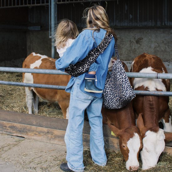 Een vrouw met een spijkerblouse en spijkerbroek draagt een baby. De draagzak heeft een luipaardprint.