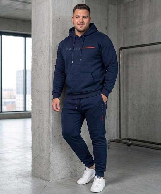 Man met donkerblauwe trainingsset: hoodie met "PARIS" opdruk en joggingbroek. In een betonnen ruimte.
