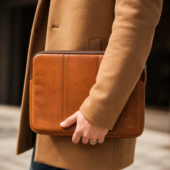 Une main avec des bagues tient une housse d'ordinateur portable marron en cuir végétalien contre un manteau de couleur camel.