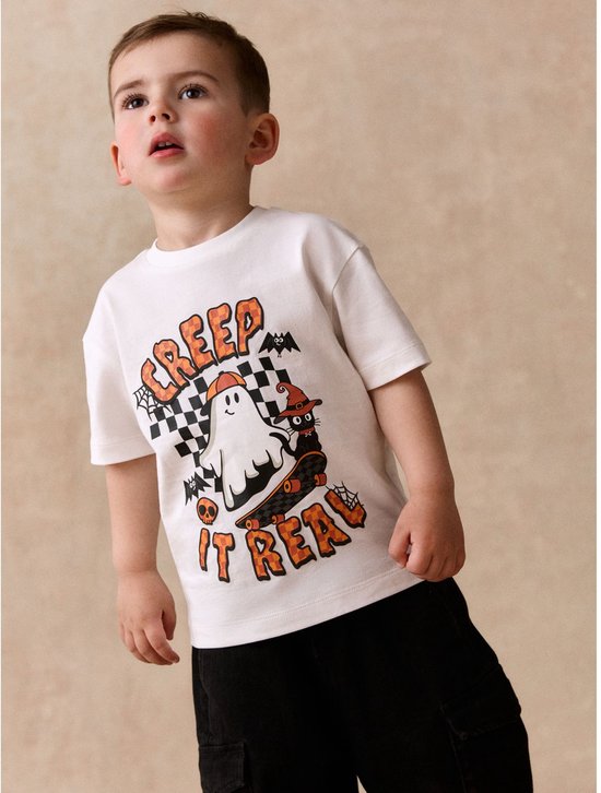 Jongen draagt een wit T-shirt met een Halloween-print van een spook op een skateboard en 'CREEP IT REAL'-tekst.
