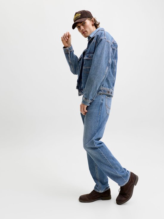 Man in een denim spijkerjasje en rechte jeans, met een bruine pet en schoenen. Staat in een modieuze pose.