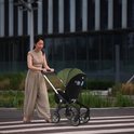Vrouw met zandkleurige broek en beige top duwt een legergroene kinderwagen met zilveren frame op een zebrapad.