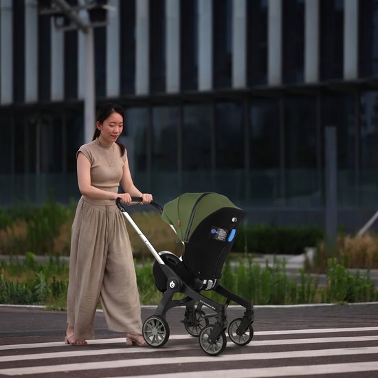 Vrouw met zandkleurige broek en beige top duwt een legergroene kinderwagen met zilveren frame op een zebrapad.