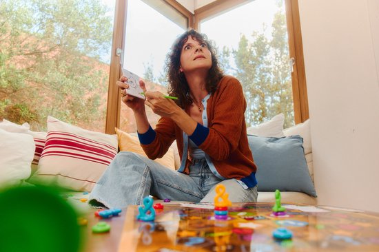 Een vrouw met bruine vest en blauwe jeans speelt een spelletje. Ze houdt een kaart omhoog en kijkt peinzend naar het plafond.