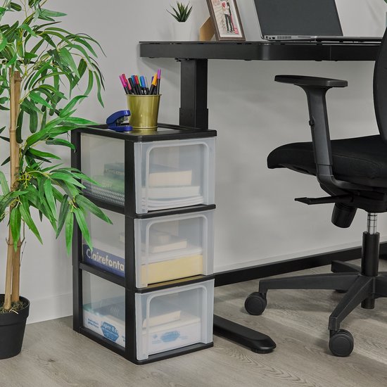 Een zwart ladesysteem met drie transparante lades, naast een bureau, met een plant.