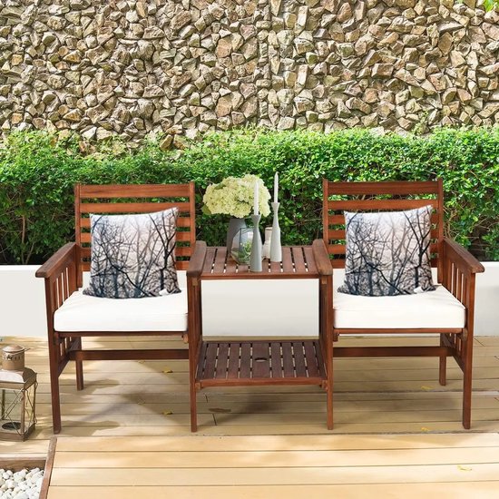 Bruine houten tuinset met witte kussens en een tafel met een afbeelding van winterse bomen op decoratieve kussens. Staat op een houten terras.