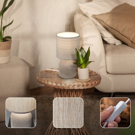 Lampe de table avec abat-jour en tissu beige et pied en céramique nervuré, sur une table d'appoint à côté d'un canapé.
