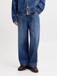 Man met korte blonde haren, donkerrode pet, jeansjack en wijde jeans, frontaal.