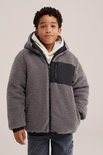 Jongen in grijze teddy jas met capuchon, beige hoodie eronder en een zwart borstvakje met rits.