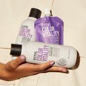 Een hand houdt een KMS Color Vitality shampoo, conditioner en gloss treatment vast. De verpakking is crèmekleurig met paarse letters.