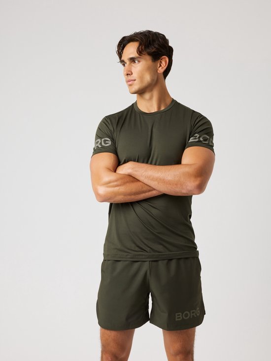 Man met gekruiste armen, draagt een donkergroen sportshirt met grijze logo opdruk op de mouw.