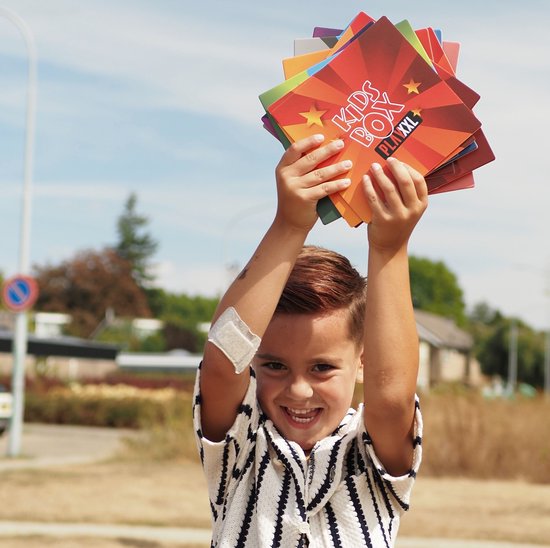 Jongen met pleister op arm houdt kleurrijke 'KIDS BOX PLAY XXL' kaarten omhoog.