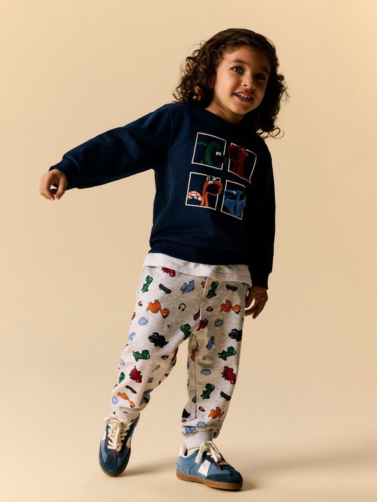 Jongetje met krullend haar draagt een donkerblauwe sweater met dinosaurussenprint en een grijze joggingbroek met dinosaurussen.